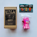 Fluorescent pink mini figurine of a rabbit, sitting with hollow blue eyes and small brown antlers coming out the side of its head, like a jackalope. It sits next to its product packaging and a postcard.