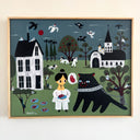 A framed folk art style painting of a woman in white holding a piece of meat next to a large black cat, with a white house, a church, trees, and birds in a stylized, muted color palette in the background.