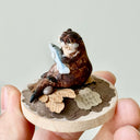 Small figurine of an otter holding a fish on a wooden base with a plain background