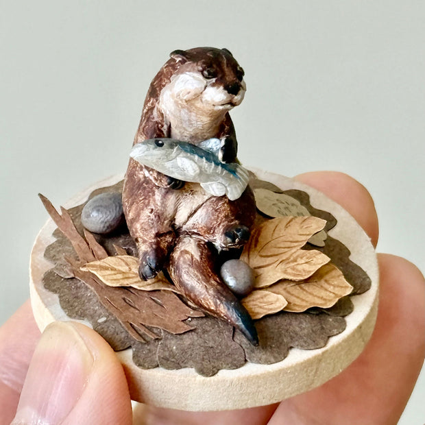 Miniature sculpture of an otter holding a fish on a wooden base with leaves.