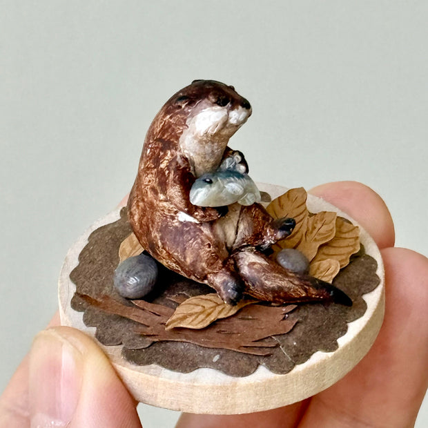 Small sculpture of 2 otters - a larger one and a baby - on a wooden base covered in paper with small leaves.