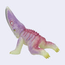 A small plastic kaiju toy figurine with a curved snout, featuring a translucent lavender-purple body, pink spikes along its back, and pale yellow feet and underbelly, set against a plain light gray background.