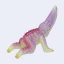 A small plastic kaiju toy figurine with a curved snout, featuring a translucent lavender-purple body, pink spikes along its back, and pale yellow feet and underbelly, set against a plain light gray background.