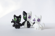 Three cat shaped figurines, with cute, large anime style eyes standing on a wooden tray with fake grass. They all have 2 tails.