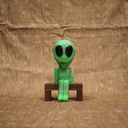 Whittled wooden sculpture of a green alien with a large head and black eyes. It has a slight frown and sits on a bench.