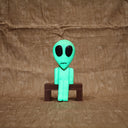 Whittled wooden sculpture of a green alien with a large head and black eyes. It has a slight frown and sits on a bench. It glows under blacklight.