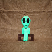 Whittled wooden sculpture of a green alien with a large head and black eyes. It has a slight frown and sits on a bench. It glows under blacklight.