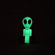 Whittled wooden sculpture of a green alien with a large head and black eyes. It has a slight frown and sits on a bench. It is shown in the dark, glowing.