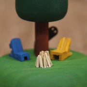 Sculptural piece of a round tree on a mound of grass. In front are 2 wooden chairs and an unlit bonfire. Behind the tree is a shadowy figure, peeking out.