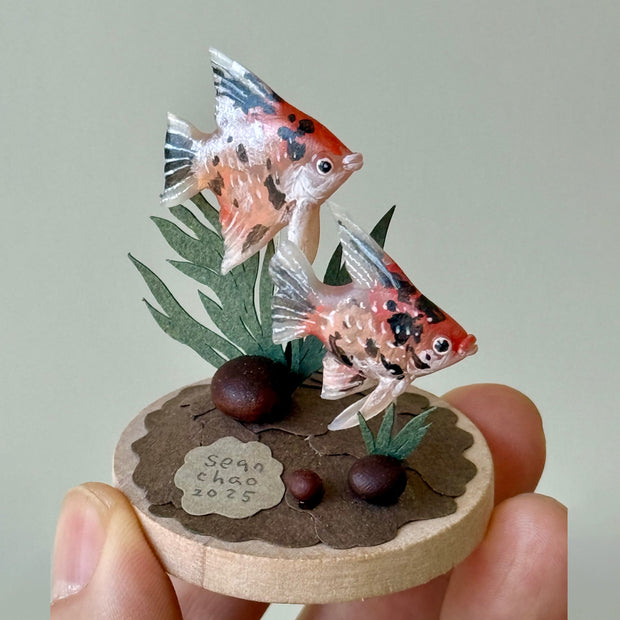 Miniature diorama with two goldfish on a circular base held by a hand.