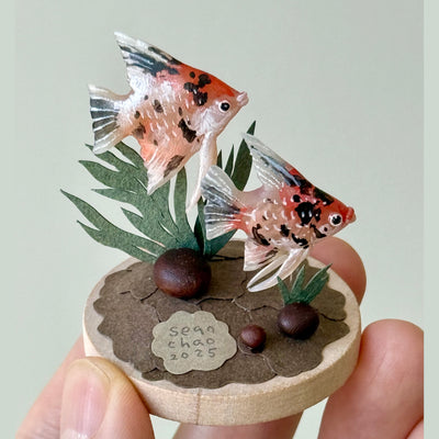 Miniature diorama with two fish on a circular base held by a hand against a light gray background