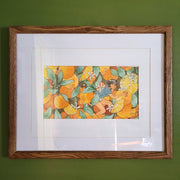 Brightly colored watercolor illustration of a girl in shorts and a tshirt sitting amongst large oranges, still on the branches. There are white orange blossoms and some open slices of the fruit. Piece is matted into a thin wooden frame.