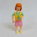 Clay figure of a woman with short orange hair and bangs, she holds a small tray with a green cup of tea on it and has her tongue sticking out. She wears a short sleeve button up, vest and skirt.