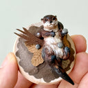Small sculpture of an otters on their back holding tiny stones on a wooden base covered in paper with small leaves.