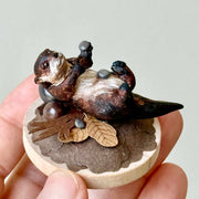 Small sculpture of an otters on their back holding tiny stones on a wooden base covered in paper with small leaves.
