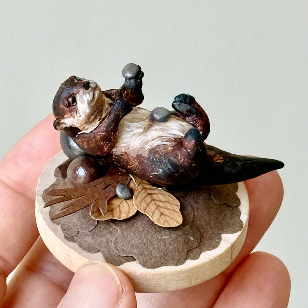Small sculpture of an otters on their back holding tiny stones on a wooden base covered in paper with small leaves.