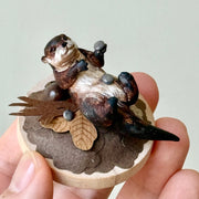 Small sculpture of an otters on their back holding tiny stones on a wooden base covered in paper with small leaves.