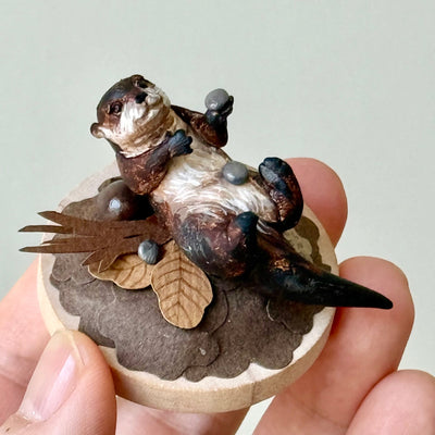 Small sculpture of an otters on their back holding tiny stones on a wooden base covered in paper with small leaves.