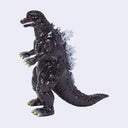 A Godzilla toy figure in a standing pose, featuring a glossy black textured body, white spiky back, and green-tipped claws and feet, set against a plain light gray background.