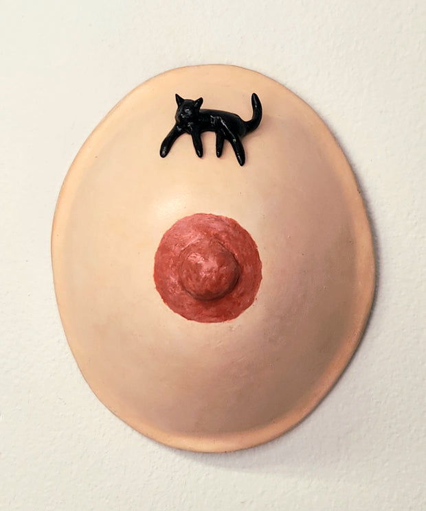 A sculpture of a single boob with a painted nipple. A small black cat rests atop the curve of the boob.