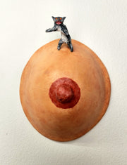 Sculpture of a single boob with a painted nipple. A small sculpted gray tabby cat stands atop it on its hind legs, with a fierce expression.