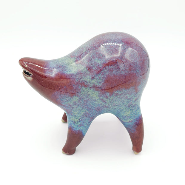 Light burgundy and blue ceramic sculpture of a rounded body quadruped creature with an open mouth goofy smile. It has small golden eyes and a dripping texture on its body.