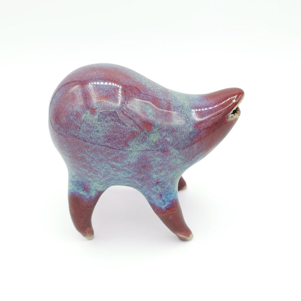 Light burgundy and blue ceramic sculpture of a rounded body quadruped creature with an open mouth goofy smile. It has small golden eyes and a dripping texture on its body.