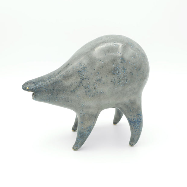 Gray ceramic sculpture of a rounded body quadruped creature with an open mouth goofy smile. It has tiny golden dot eyes blue speckled body.