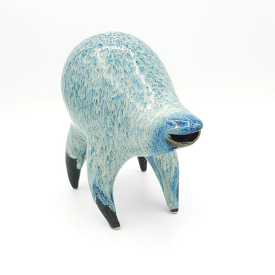 Blue and white ceramic sculpture of a rounded body quadruped creature with an open mouth goofy smile. It has small golden eyes and a dripping texture on its body.