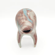 Blue and dusky pink ceramic sculpture of a rounded body quadruped creature with an open mouth goofy smile. It has small golden eyes and a swirled texture on its body.
