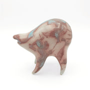Blue and dusky pink ceramic sculpture of a rounded body quadruped creature with an open mouth goofy smile. It has small golden eyes and a swirled texture on its body.