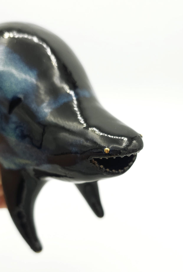 Black and blue ceramic sculpture of a rounded body quadruped creature with an open mouth goofy smile. It has tiny golden dot eyes and a galactic pattern body.