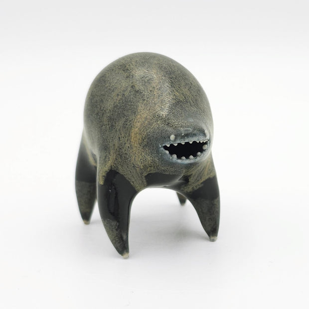 Black and dark gray ceramic sculpture of a rounded body quadruped creature with an open mouth goofy smile. It has small silver eyes and a gritty texture on its body.
