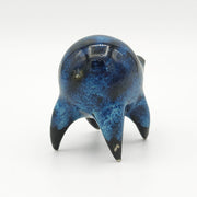 Black and blue ceramic sculpture of a rounded body quadruped creature with an open mouth goofy smile. It has small golden eyes and a swirling texture on its body.