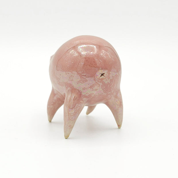Light pink ceramic sculpture of a rounded body quadruped creature with an open mouth goofy smile. It has small golden eyes and an oil slick texture on its body.