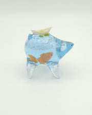 Resin sculpture of a blue rounded body quadruped creature with the illusion of water inside of its body. A goldfish swims at the bottom of its stomach and atop its back is a paper boat.