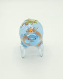 Resin sculpture of a blue rounded body quadruped creature with the illusion of water inside of its body. Several goldfish swim within its stomach and atop its back is a small paper boat.