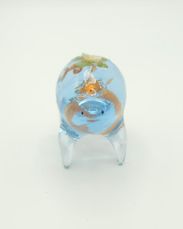 Resin sculpture of a blue rounded body quadruped creature with the illusion of water inside of its body. Several goldfish swim within its stomach and atop its back is a small paper boat.