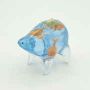 Resin sculpture of a blue rounded body quadruped creature with the illusion of water inside of its body. Several goldfish swim within its stomach and atop its back is a small paper boat.