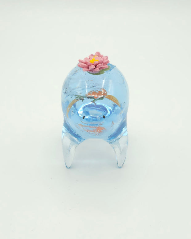 Resin sculpture of a blue rounded body quadruped creature with the illusion of water inside of its body. A large goldfish swims within its stomach and atop its back are 2 pink water lilies.