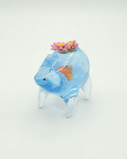 Resin sculpture of a blue rounded body quadruped creature with the illusion of water inside of its body. A large goldfish swims within its stomach and atop its back are 2 pink water lilies.