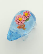 Resin sculpture of a blue rounded body quadruped creature with the illusion of water inside of its body. A large goldfish swims within its stomach and atop its back are 2 pink water lilies.