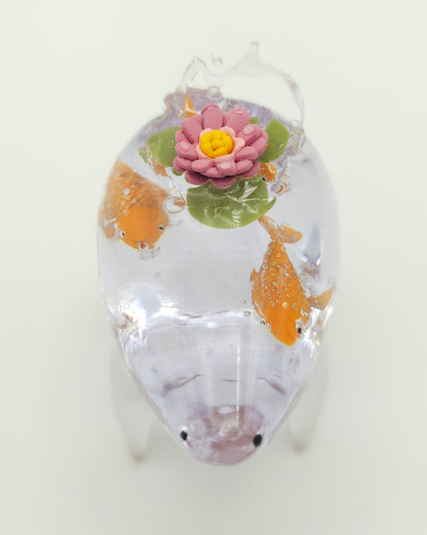 Resin sculpture of a clear rounded body quadruped creature with the illusion of water inside of its body. Several goldfish swim within its stomach and atop its back is a pink water lily with lily pads.