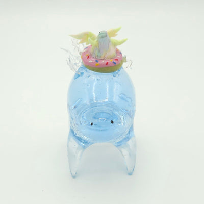 Resin sculpture of a blue rounded body quadruped creature with the illusion of water inside of its body. On its back is a pastel winged chubby creature in a donut shaped inflatable tube.