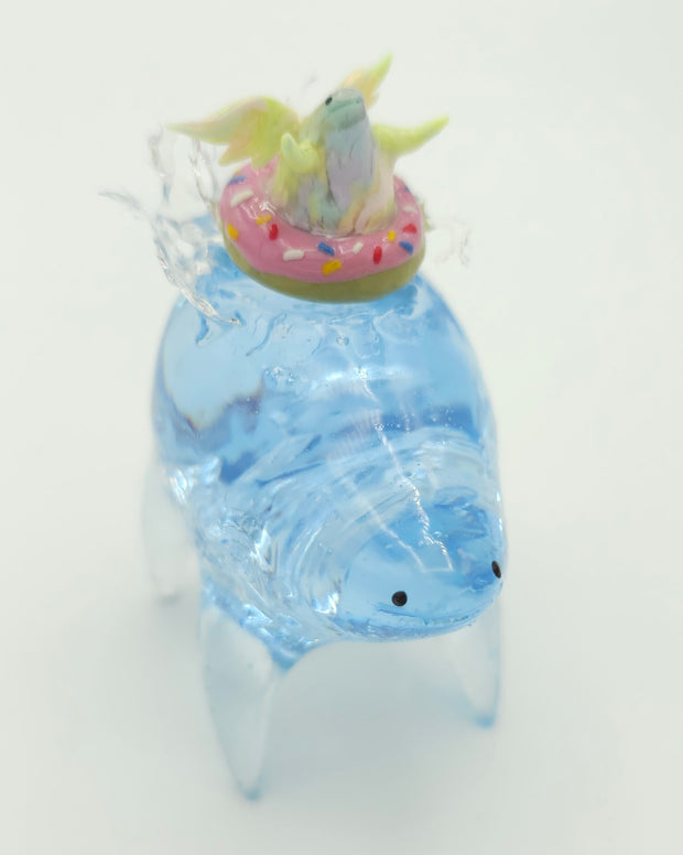 Resin sculpture of a blue rounded body quadruped creature with the illusion of water inside of its body. On its back is a pastel winged chubby creature in a donut shaped inflatable tube.