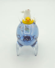 Resin sculpture of a blue rounded body quadruped creature with a goldfish swimming inside its body. On its back is a white winged chubby creature in a rubber duck shaped inflatable tube.