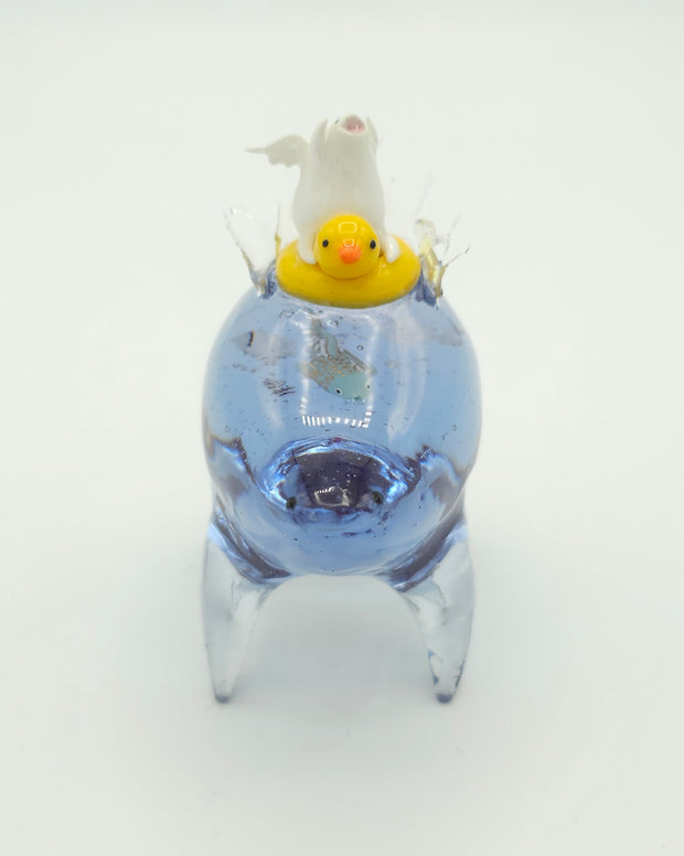 Resin sculpture of a blue rounded body quadruped creature with a goldfish swimming inside its body. On its back is a white winged chubby creature in a rubber duck shaped inflatable tube.