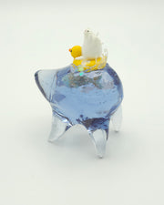 Resin sculpture of a blue rounded body quadruped creature with a goldfish swimming inside its body. On its back is a white winged chubby creature in a rubber duck shaped inflatable tube.