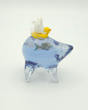 Resin sculpture of a blue rounded body quadruped creature with a goldfish swimming inside its body. On its back is a white winged chubby creature in a rubber duck shaped inflatable tube.