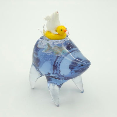 Resin sculpture of a blue rounded body quadruped creature with a goldfish swimming inside its body. On its back is a white winged chubby creature in a rubber duck shaped inflatable tube.